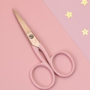 May include: Pink metal scissors with a gold screw and the words "SUPER ART" on the handle. The scissors are on a pink background with gold stars.