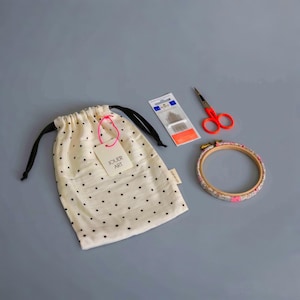 Puede incluir: Una bolsa de cordón de algodón blanco con lunares negros y una etiqueta que dice "Jouer Art". La bolsa está al lado de un pequeño paquete de agujas de bordado, un par de tijeras rojas y un aro de bordado floral.