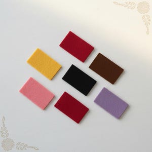 Peut inclure: Sept échantillons de feutrine rectangulaires disposés en groupe. Les couleurs incluent le rouge, le jaune, le rose, le rouge foncé, le noir, le marron et le violet clair. Chaque pièce présente une texture uniforme et une forme identique.