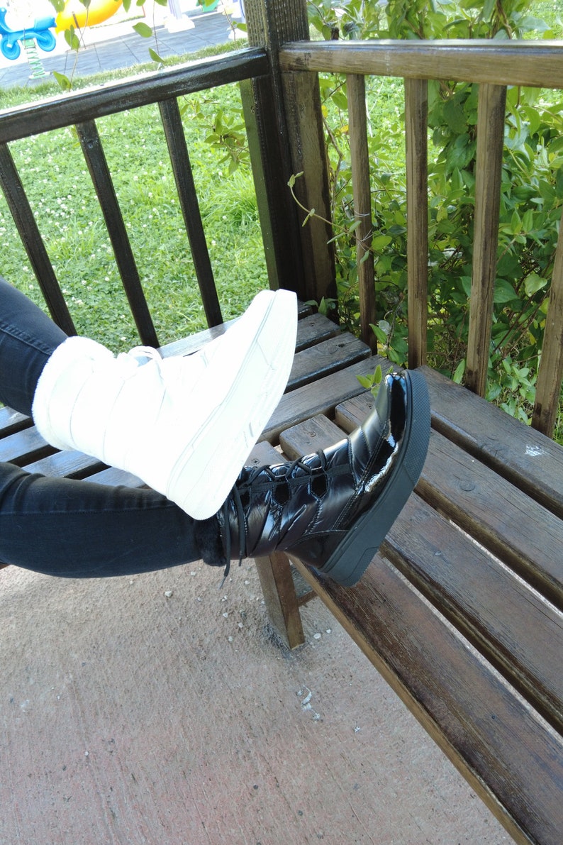 May include: A pair of black and white boots with laces. The black boot is on the right foot and the white boot is on the left foot. The boots are worn by a person sitting on a wooden bench.