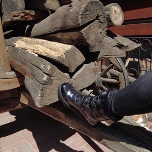 May include: A black leather boot with a zipper on the side is resting on a pile of stacked wooden logs. The boot is on a wooden cart with a wheel visible in the background.