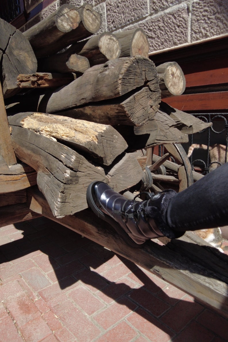 May include: A black leather boot with a zipper on the side is resting on a pile of stacked wooden logs. The boot is on a wooden cart with a wheel visible in the background.