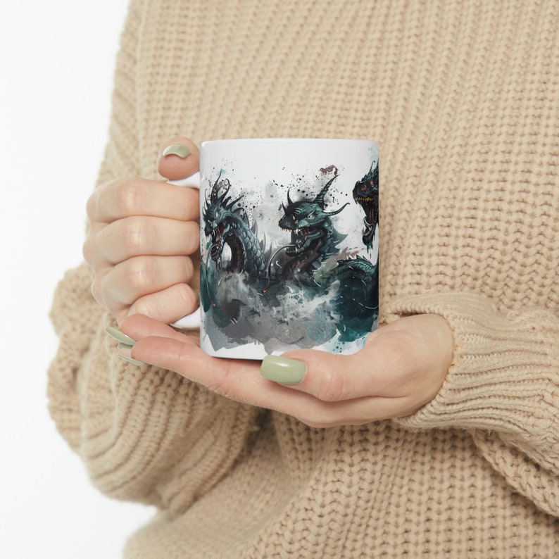 May include: White ceramic mug featuring a detailed illustration of three dragons in shades of teal, black, and gray. The dragons are depicted with intricate scales and horns, set against a misty, watercolor-style background. The mug has a standard handle.
