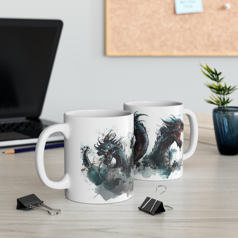 May include: Two white ceramic mugs with a dragon design. The dragon is depicted in shades of black, teal, and red, with a watercolor effect. The mugs are on a light-colored wooden surface, with a laptop and office supplies in the background.