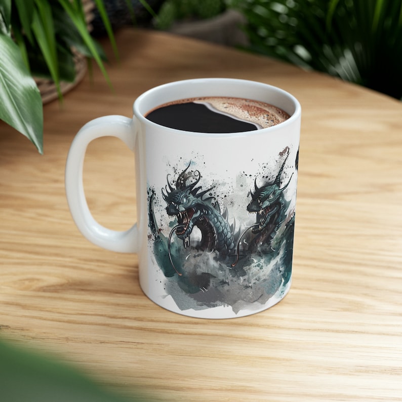 May include: White ceramic coffee mug featuring a detailed illustration of two teal dragons with open mouths, set against a watercolor-style background. The mug is filled with dark coffee and has a white handle.