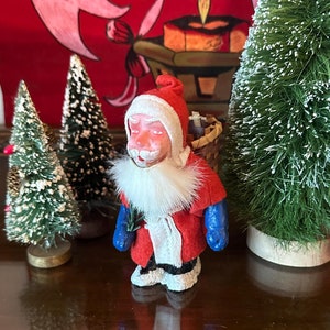May include: A vintage Santa Claus figurine with a red and white suit, blue mittens, and a white beard. He is carrying a basket on his back and is standing in front of two small artificial pine trees.