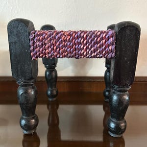 May include: A small wooden stool with four turned legs and a woven fabric seat in shades of purple, pink, and red.