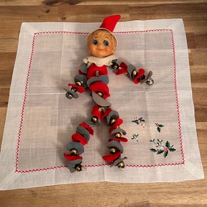 May include: A vintage elf doll with a red hat and a gray and red body made of fabric circles. The doll is lying on a white linen handkerchief with red embroidered edging.