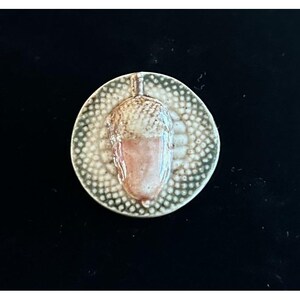 May include: A ceramic acorn pendant with a textured, speckled background. The acorn is light brown and pink, with a stem at the top. The pendant is circular and has a rustic, handmade appearance.