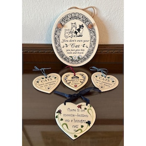 May include: A collection of cat-themed ceramic decorations. Includes an oval plaque with cat illustrations and text, and heart-shaped ornaments with cat-related quotes. The color palette is primarily cream, black, and brown.