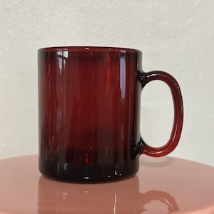 May include: A ruby red glass mug with a rounded handle. The mug has a cylindrical shape and a smooth, glossy finish. The mug is sitting on a pink surface.