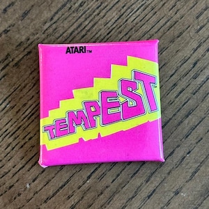 May include: A square pinback button featuring a bright pink background with a diagonal yellow stripe. The word TEMPEST is printed in large pink letters across the yellow stripe, and the word ATARI is printed in small black text at the top.