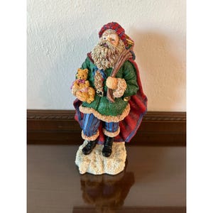 May include: A Santa Claus figurine with a long white beard, wearing a green coat with red trim and a red cape. He holds a teddy bear and a bundle of sticks. The figurine stands on a white base.