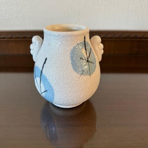 May include: A small, off-white ceramic vase with a textured, pebbled surface. The vase features two small, wing-shaped handles on the sides and is decorated with light blue circular patches accented by thin, black, hand-painted linear markings.