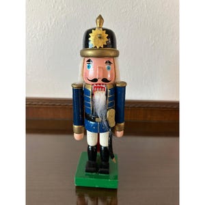 May include: A wooden nutcracker figurine wearing a black helmet with a gold star emblem, a blue jacket with gold trim, white trousers, and black boots. The figure has a white beard, painted blue eyes, and a black mustache, standing on a green base.