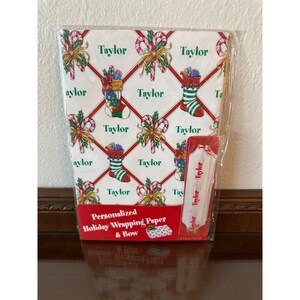 May include: Personalized holiday wrapping paper and bow set. The wrapping paper features a repeating pattern of Christmas stockings, candy canes, and bows on a white background. The name "Taylor" is printed throughout the design. The set includes a matching gift tag.