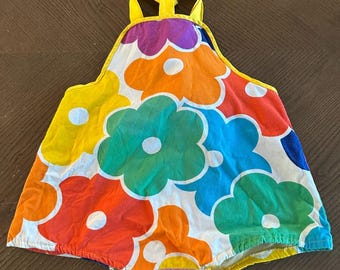 May include: A colorful romper with a vibrant floral pattern. The romper features large flowers in shades of red, orange, yellow, green, blue, and purple against a white background. It has yellow straps and trim.