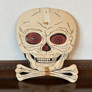 May include: A vintage die-cut cardboard skull and crossbones decoration in cream, black, and dark red. The skull features deep-set red eyes with black circular pupils, a black nose cavity, and a wide grin. The text MADE IN U.S.A. is printed on the bone.