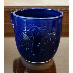 May include: A cobalt blue ceramic mug with a bird design. The mug has a rounded shape with a handle and a white base. The bird design is etched into the surface of the mug.