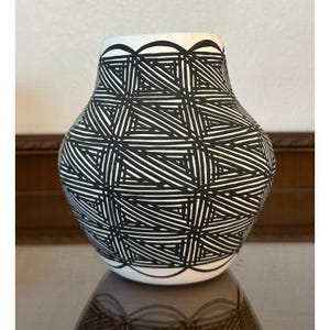 May include: A white ceramic vase with a black geometric pattern. The vase has a rounded body and a slightly flared rim. The black pattern consists of intersecting lines, creating a textured effect. The rim and base feature a scalloped design.