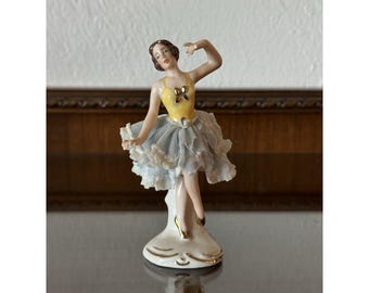 May include: A porcelain figurine of a ballerina in a yellow bodice and a light blue tutu with lace trim. The figurine has brown hair and is posed mid-dance. Gold accents adorn the base and ballet slippers. A decorative item.