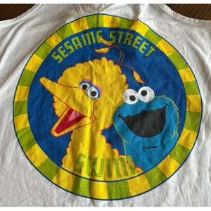 May include: White t-shirt featuring a circular graphic with Big Bird and Cookie Monster from Sesame Street. The graphic is in yellow, blue, and green, with the words "SESAME STREET" and "SWIM" visible.