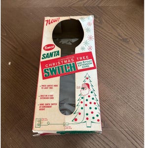May include: A rectangular cardboard box for a Timco Santa Combination Christmas Tree Switch with an 8 foot extension cord. The box features red and green text, a graphic of a Christmas tree, and a circular cutout for the Santa-shaped switch.