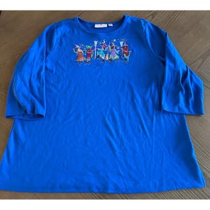 May include: A long-sleeved, royal blue shirt with an embroidered design of several figures in colorful costumes. The figures are in a line, with hats and accessories. The shirt has a crew neck and three-quarter sleeves.