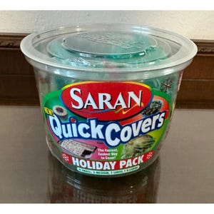 May include: A clear plastic container with a green lid, labeled "SARAN Quick Covers Holiday Pack." The label features red, green, and white text and images of food items. The pack includes various sizes of covers.