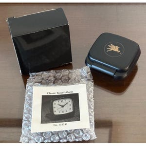 May include: A black square travel alarm clock case with a gold Pegasus logo on the lid, a black cardboard box, and a white instruction card labeled Classic Travel Alarm No. 122741, all packaged with a clear plastic bubble wrap protective sheet.