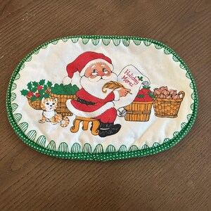 May include: An oval fabric placemat with a white background and a green polka-dot border. The center features a graphic of Santa Claus sitting on a stool, holding a quill and a scroll labeled Holiday Menu, with a cat and baskets of produce.
