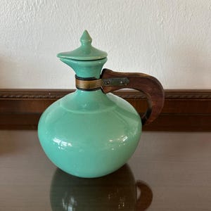 May include: A mint green ceramic pitcher with a rounded body and a pointed lid. The pitcher features a dark brown wooden handle attached to the neck with a metallic band. The surface of the ceramic is smooth and glossy, reflecting light.