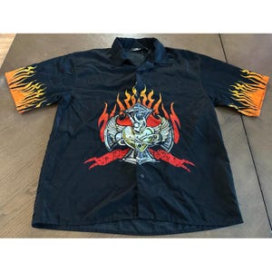 May include: Black button-down shirt with a graphic design featuring a heart, wings, and a cross, with flames on the sleeves. The graphic is in shades of red, yellow, and white.
