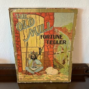 May include: A vintage board game box titled The Old Mill Fortune Teller. The cover features a colorful illustration of a person with a beard and hat sitting before a wooden door and stone wall, with a waterfall and text printed in black.