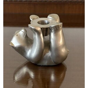 May include: A silver-colored bear-shaped candle holder. The bear is depicted in a playful pose, holding a circular candle cup. The metal has a polished finish, reflecting light. The candle holder is designed to hold a single candle.