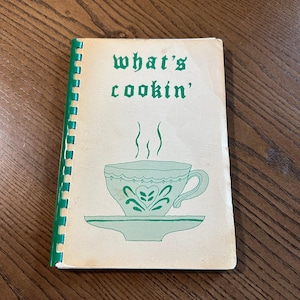May include: A spiral-bound cookbook with a cream-colored cover featuring the green text what's cookin' and a green illustration of a teacup on a saucer with steam rising from the top. The binding is a bright green plastic coil.