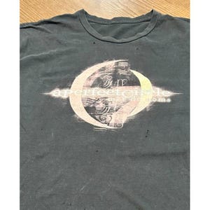 May include: Dark gray t-shirt featuring a circular design with the text "a perfect circle" and "mer de noms" in a light beige color. The shirt has a crew neck and appears worn.