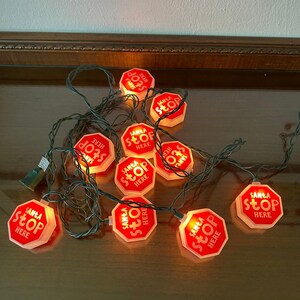 May include: A string of holiday lights featuring ten octagonal red and white plastic signs. Each sign is illuminated from within and displays the text SANTA STOP HERE in white lettering. The lights are connected by a dark green electrical cord.