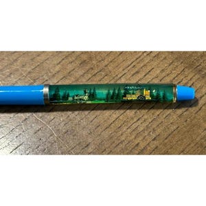 May include: A blue ballpoint pen featuring a clear, liquid-filled barrel section that displays a miniature scene of a steam locomotive train traveling through a dense green forest of pine trees, with a yellow train car visible on the right side.