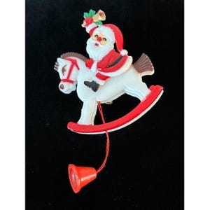 May include: A Christmas-themed plastic ornament featuring Santa Claus riding a white rocking horse with red accents. Santa wears a red suit and hat, holding a bell. A red string connects to a red bell below.
