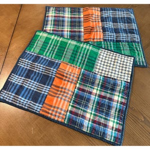 May include: Two square patchwork placemats with a variety of plaid patterns in blue, green, orange, and white. The placemats are quilted with a dark blue border and are displayed on a wooden table.