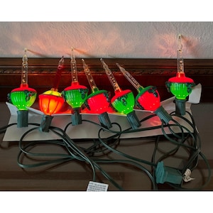 May include: A string of bubble Christmas lights featuring clear glass tubes filled with liquid, set atop colorful bases in shades of red and green. Each light is shaped like a decorative ornament with holly leaf accents, connected by a dark green wire.