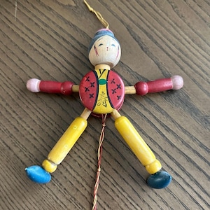 May include: A wooden jumping jack toy featuring a painted figure with a round torso, red arms, yellow legs, and blue feet. The torso displays a red and yellow pattern with black markings and a central character. A string hangs from the bottom.