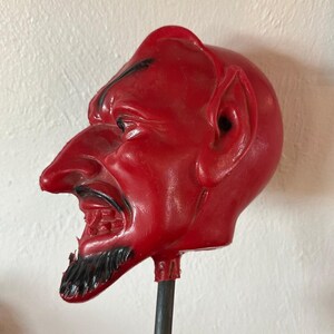 May include: A red molded wax candle shaped like a devil head in profile. The figure features a pointed nose, a sharp chin, and a black painted goatee and eyebrow. The candle is mounted on a thin, dark metal rod, showing a smooth, glossy finish.