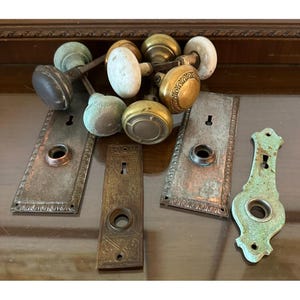 May include: A collection of antique door hardware, including various doorknobs and matching backplates. The doorknobs are made of brass, porcelain, and other materials, with a range of colors from gold to white and green. The backplates are rectangular and ornate.