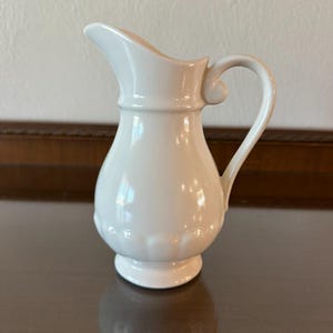 May include: A white ceramic mug with a black handle and the text "But First, Coffee" in a bold, black font. The mug is set against a blurred background, suggesting a cozy setting.