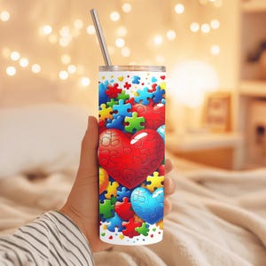 May include: A white tumbler with a stainless steel straw, featuring a design of colorful puzzle pieces and heart shapes. The tumbler is cylindrical and appears to be approximately 8 inches tall.