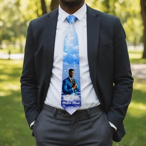 May include: A blue tie with a sky and cloud design, featuring a photo of a man in a blue suit. The tie also has a white dove and the name "Milton Mane" printed on it. The tie is worn with a white shirt and a black suit.