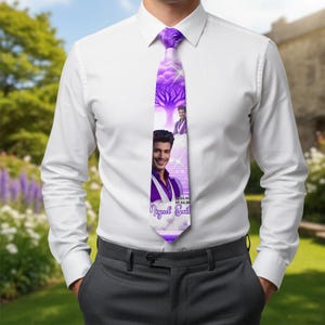 May include: A purple tie featuring a portrait of a man, a tree design, and the name "Miguel Sani" in white script. The tie is worn with a white dress shirt and gray pants. The tie's design includes a date.