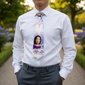 May include: A white tie featuring a portrait of a woman in a purple top, with a white dove and a light purple floral design. The tie is worn with a white dress shirt and gray pants.